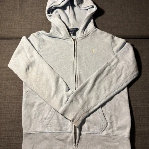 Ljusblå hoodie från Polo Ralph Lauren - Ljusblå fläckfri hoodie från Polo Ralph Lauren med dragkedja, huva och klassisk broderad logga på bröstet.Tröjan har känguruficka och är tillverkad i mjuk bomull. Tröjan är mer blå i verkligheten och har använts få antal gånger. Den är även rätt runn så passar bra till sommar, storlek 152. Pris kan diskuteras.