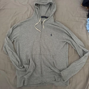Grå hoodie från Polo Ralph Lauren - Säljer en riktigt fet grå hoodie från Polo Ralph Lauren med dragkedja och huva. Klassisk broderad logga på bröstet, vita snören och två fickor framtill. Tillverkad i mjuk bomullsblandning, perfekt för chill dagar. Långärmad och normal passform. Om du vill ha fler bilder eller fråga något så tveka inte att fråga ! 