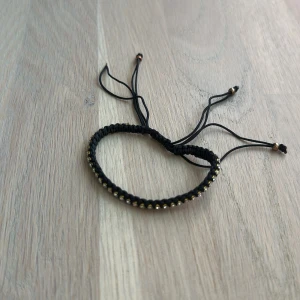 Svart flätat armband med gulddetaljer - Snyggt svart armband med flätad design och små guldfärgade pärlor längs hela bandet. Justerbar knytning med tunna snören och små pärlor i ändarna. Perfekt accessoar för en cool och stilren look.