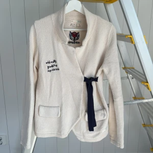 Beige cardigan jacka från Odd Molly - Superfin beige cardigan jacka från Odd Molly med broderad ros på ryggen och bröstet. Jackan har omlottmodell med mörkblå knytband i midjan, två fickor fram och coola broderade detaljer. Perfekt för dig som gillar unika plagg med snygga detaljer.