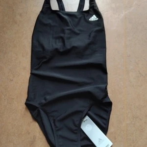 Helt Ny Adidas Baddräkt - En svart baddräkt från Adidas, helt ny med alla lappar kvar. Logga på ena bröstet, snygg öppen rygg, samt liten text "Primeblue" i ryggslutet. Storlek XS men sitter som S. Köpt för 399 kr. 