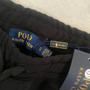 Svarta shorts från Polo Ralph Lauren - Svarta shorts från Polo Ralph Lauren med klassisk vit logga broderad på benet. Mjukt bomullsmaterial, elastisk midja med snörning och två sidofickor. Perfekta för chill dagar eller när du vill ha en clean och stilren look.