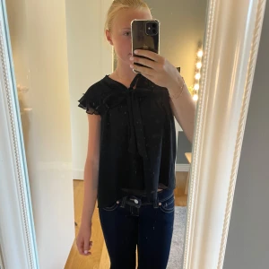 Svart blus med volang och rosett - Supersöt svart blus med volangdetaljer vid axlarna och stor rosett framtill vid halsen. Ärmlös modell i ett lätt och flowy tyg som ger en luftig känsla. Perfekt att styla med jeans eller kjol för en trendig look.