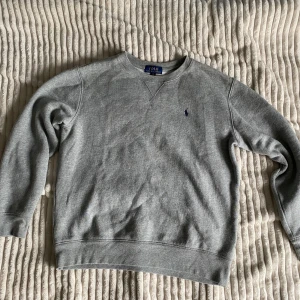 Grå sweatshirt från Polo Ralph Lauren - Klassisk grå sweatshirt från Polo Ralph Lauren med rund halsringning och broderad marinblå logga på bröstet. Tröjan har långa ärmar och ribbade muddar vid ärmslut och nederkant. Perfekt för en chill och stilren look. Kommer från ett hem med katt. Hör av dig om du har några frågor! 