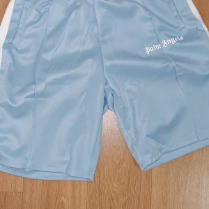 Palm Angels ljusblå shorts - Snygga ljusblå shorts från Palm Angels med vit logga tryckt på ena benet. Shortsen har vita paneler på sidorna och är gjorda i ett glansigt syntetmaterial. Perfekta för en sportig look eller chill vardagsstil.