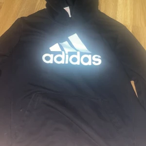Svart Adidas hoodie med stor logga - Svart hoodie från Adidas med stor vit logga på bröstet. Klassisk känguruficka framtill och huva. Perfekt för dig som gillar sportig och avslappnad stil. Mjuk och skön, passar till chill eller träning.