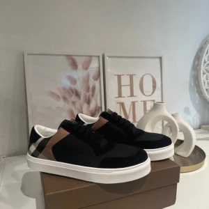 Burberry sneakers svart/vit - Snygga Burberry sneakers med klassiskt rutigt mönster i svart, beige och vitt på hälen. Ovandel i svart textil och mocka, vita sulor och svarta skosnören. Perfekta för dig som vill ha en stilren och lyxig look med ikoniska Burberry-detaljer. Kvitto finns!
