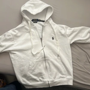 Vit hoodie från Polo Ralph Lauren bra skick nästan helt ny  - Säljer en clean vit hoodie från Polo Ralph Lauren med dragkedja, snörning i huvan och klassisk logga på bröstet. Tröjan har känguruficka och är i mjuk bomull, perfekt för chill dagar. Custom fit-modell för snygg passform.