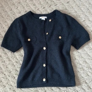 Mörkblå stickad tröja med guldknappar - Snygg mörkblå stickad kofta med korta ärmar och guldiga knappar framtill. Koftan har två små fickor på bröstet och ribbade kanter vid ärmslut och nederkant. Använd fåtal gånger. Pris kan diskuteras 🤍
