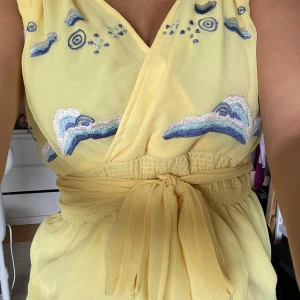 Gul blus - Superfin gul omlottblud med broderade blå och vita moln och detaljer upptill. Klänningen har knyt i midjan och är ärmlös, perfekt för en somrig look. Lätt och luftig känsla, passar dig som vill sticka ut med coola detaljer. Är i princip i nyskick.
