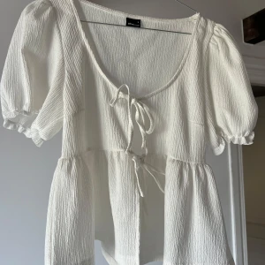 Vit blus med knyt från Gina Tricot - Supersöt vit blus från Gina Tricot med puffiga korta ärmar och två knyt framtill. Blusen har en lätt rynkad struktur och volang nedtill. Djup rundringning och luftig passform. 