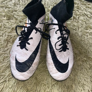Nike Hypervenom vita fotbollsskor - Nike Hypervenom fotbollsskor i vitt med svarta detaljer och stora svarta swoosh-loggor. Skorna har högt skaft i svart, snörning och färgglada mönster i gult, orange och rosa längs sidorna. Sulan är gul med orange dobbar. Perfekta för dig som vill sticka ut på planen. Finns ett hål i strumpan 