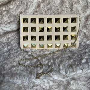 Beige kuvertväska med guldiga nitar - Kuvertväska i beige mocka med stora fyrkantiga guldnitar på framsidan och en tunn guldfärgad kedja som axelrem. Väskan har en stilren och edgy look, perfekt för att lyfta din outfit. Ingen synlig logga eller märke. Fodret har väldigt mycket sminkfläckar.