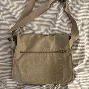 Beige axelväska från Björn Borg - Säljer en beige axelväska från Björn Borg med justerbar rem och flera praktiska fack. Väskan har dragkedja framtill, svart detalj och tryckt logga. Rymmer laptop och annat viktigt. Lite sliten men påverksr inte funktionen eller något på nå sätt:)