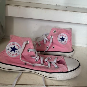Rosa Converse All Star sneakers - Klassiska Converse All Star sneakers i rosa canvas med vita snören och gummisula. Hög modell med rund tå och ikonisk logga på sidan. Perfekta för en avslappnad och cool stil.