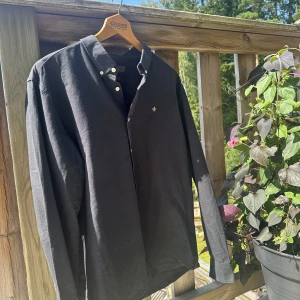 Morris skjorta - Svart långärmad skjorta från Morris i storlek XXL men som snarare sitter som en XL/L. Skjortan har klassisk button down-krage, vita knappar och en liten broderad logga på bröstet. Tillverkad i mjuk bomull och passar perfekt till dig som gillar stilren och enkel design.