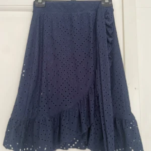 Mörkblå volangkjol från Vero Moda - Supersöt mörkblå kjol från Vero Moda med broderade hålmönster och volangkant nertill. Kjolen har omlottkänsla och knytband i sidan, perfekt för dig som gillar en feminin och trendig look. Passar extra bra till vår och sommar!