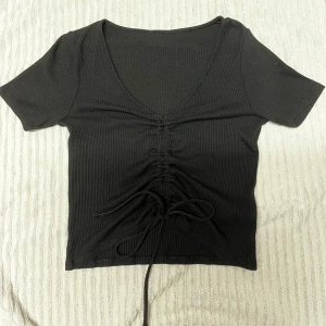 Svart ribbad topp med snörning - Svart ribbad topp med korta ärmar och v-ringning. Toppen har snygg snörning framtill som ger en cool rynkad effekt och croppad passform. Perfekt att styla med jeans eller kjol för en trendig look.