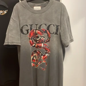 Grå Gucci t-shirt med ormtryck - Snygg grå t-shirt från Gucci med stort svart logotryck och en röd orm på framsidan. Oversized fit och klassisk rund hals. Tillverkad i mjuk bomull som känns skön mot huden. Perfekt statement-plagg för dig som vill sticka ut. Använd 6 gånger