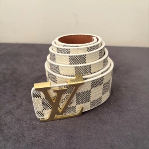 Rutig Louis Vuitton bälte med LV-spänne - Snyggt bälte från Louis Vuitton med klassiskt rutigt mönster i vitt och grått. Bältet har ett stort guldfärgat LV-spänne och är gjort i skinn. Perfekt accessoar för att lyfta din outfit med en exklusiv touch.