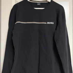 Svart långärmad tunn pyjamas tröja från BOSS - Svart långärmad tunn pyjamas tröja från BOSS i bomull med stilren design. Framtill finns en tunn vit och beige linje samt BOSS-logga tryckt på bröstet. Tröjan har rund halsringning och en klassisk passform.