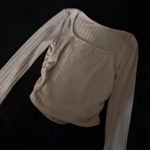 Beige ribbad långärmad topp - Trendig beige topp med ribbad struktur och lång ärm. Toppen har en snygg rynkdetalj på sidan och en fyrkantig ringning som ger en modern vibe. Perfekt att styla med jeans eller kjol för en cool look.