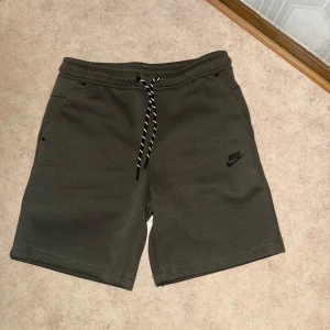 Nike tech fleece shorts - Snygga gröna Nike shorts med svart logga på benet. De har elastisk midja med svartvit snörning och praktiska fickor, varav en med dragkedja på sidan. Perfekta för chill eller träning, i mjukt bomullsmaterial och relaxed fit.