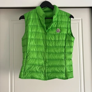 Grön dunväst från Moncler - Snygg grön dunväst från Moncler med klassisk quiltad design och tryckknappar framtill. Västen har hög krage och ikonisk Moncler-logga på bröstet. Perfekt att slänga över en hoodie eller tröja för en cool look. Näst intill aldrig använd
