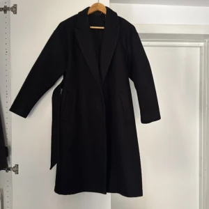Svart kappa från Vero Moda, stl S - Stilren svart kappa från Vero Moda i klassisk modell med bälte i midjan och två fickor framtill. Kappan har lång ärm och är tillverkad i en ullblandning som håller dig varm under kyliga dagar. Perfekt för dig som gillar en clean och tidlös look. Jackan är lite nopprig, samt har ett litet hål inne i vänster ficka, annars är den hel och ren.