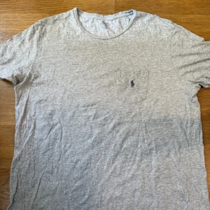 Grå t-shirt från Polo Ralph Lauren XL - Snygg grå t-shirt från Polo Ralph Lauren i storlek XL. Klassisk modell med rund hals, korta ärmar och liten bröstficka med broderad logga. Tillverkad i mjuk bomull som är skön att ha på sig hela dagen.