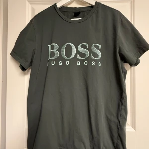 Svart t-shirt från Hugo Boss - Snygg mossgrön t-shirt från Hugo Boss med stort BOSS-tryck på bröstet. Klassisk rund halsringning och korta ärmar. Tillverkad i mjuk bomull som känns skön mot huden. Perfekt för dig som gillar stilrena och trendiga plagg med tydlig logga.