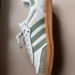 Adidas Samba vita sneakers med gröna ränder - Adidas Samba sneakers i vitt skinn med gröna ränder på sidorna och gråa mockadetaljer vid tån. Nypris 1200! Skorna är andvända ett fåtal gånger. KÖPAREN STÅR FÖR FRAKT