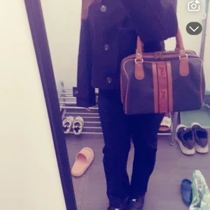 Svart och brun handväska från YSL - Säljer en snygg svart handväska från YSL med bruna detaljer och handtag. Väskan har två breda ränder i brunt och orange med YSL-logga framtill. Perfekt storlek för vardagsgrejer och ger en lyxig vibe till outfiten.