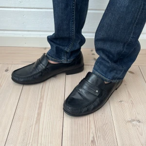 Svarta loafers från Aristokrat i skinn - Klassiska handgjorda svarta loafers från Aristokrat i äkta skinn. Snygg penny loafer-design med bred passform och diskret detalj på sidan. Perfekta till jeans eller chinos för en clean och tidlös look. 