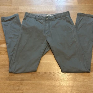 Grå Bowery chinos slim fit W30 L32 - Snygga grå chinos från Bowery i slim fit-modell. Byxorna har klassisk design med fickor fram och bak, bälteshällor och stängs med knapp och dragkedja. Perfekta för en clean och stilren look.