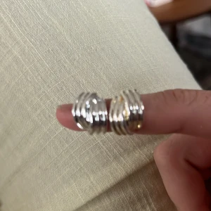 Två chunky ringar i silver och guld - Säljer två coola chunky ringar i silverfärgad metall, en av dem har även detaljer i guld. Båda ringarna har ett flerradigt, nästan virat utseende som ger en modern och edgy vibe. Perfekta för att lyfta din stil och matcha med andra smycken.