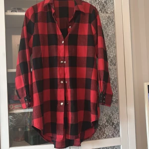 Röd och svart rutig skjorta med tryckknappar - Snygg röd och svart rutig skjorta i oversized modell med långa ärmar och silvriga tryckknappar framtill och vid ärmslut. Skjortan har klassisk krage och är tillverkad i mjuk bomull, perfekt för en avslappnad och cool stil.