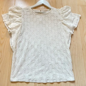 Vit blus med puffärm Holly &amp; Whyte - Supersöt vit blus från Holly &amp; Whyte med korta puffärmar och diskret strukturmönster. Blusen har rund halsringning och knapp i nacken. 💗