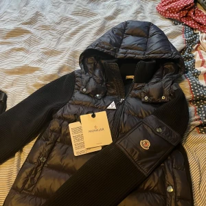 Svart Moncler pufferjacka med huva - Snygg svart pufferjacka från Moncler med stickade ärmar och vadderad kropp. Jackan har huva, dragkedja framtill och Moncler-logga på ärmen. Perfekt för kalla dagar och ger en clean, exklusiv look. Materialmix av polyester och stickat ger både värme och stil.