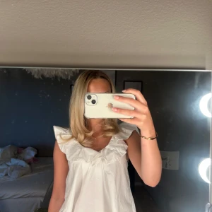 Vit blus med volangkrage - Supersöt vit blus med bred volangkrage och ärmlös design. Luftig passform och lätt material, perfekt för varma dagar. Blusen har en feminin v-ringning och volangdetaljer som ger en romantisk vibe. Snygg att styla med jeans eller kjol.