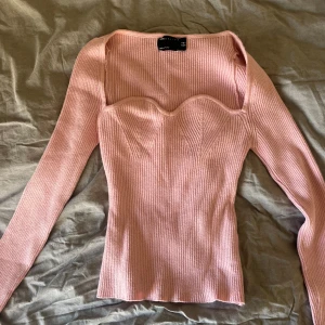 Rosa ribbad stickad topp med sweetheart - Supersöt ljusrosa stickad topp med ribbad struktur och lång ärm. Snygg sweetheart-urringning och figurnära passform som framhäver siluetten. Perfekt för dig som vill ha en trendig och feminin look.