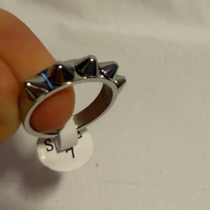 Silvrig ring med nitar, storlek 7 - Cool ring i silverfärgad metall med fem konformade nitar runt hela ringen. Perfekt för dig som gillar en edgy och alternativ stil. Ringen är i storlek 7 och har en blank finish som ger ett snyggt intryck.