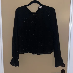 Svart glittrig blus från Gina Tricot - Supersnygg svart blus från Gina Tricot med glittriga detaljer. Blusen har långa ärmar med volang vid ärmslut och en lätt croppad passform. 