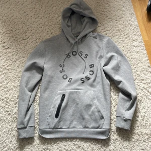 Hugo boss hoodie - Snygg grå hoodie från BOSS med svart cirkelformat BOSS-tryck på bröstet. Tröjan har huva med snörning, känguruficka med dragkedjedetalj och ribbade muddar. Perfekt för en chill och stilren look.
