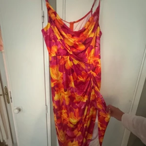 Färgstark blommig midiklänning - Säljer en maxad midiklänning med tropiskt blommönster i rosa, orange och gul. Klänningen har smala axelband, draperad framsida/baksida och snygg slits. Perfekt för sommarens alla tillfällen när du vill sticka ut.
