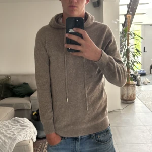 Beige stickad hoodie med snörning  (Kashmir)  - Mysig beige hoodie i stickat material med långa ärmar och huva. Tröjan har snörning i halsen och ribbade muddar vid ärmslut och nederkant. Perfekt för dig som gillar en chill och stilren look.