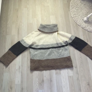 Randig polotröja från Jacqueline de Yong - Supermysig stickad polotröja från Jacqueline de Yong i storlek S. Tröjan har breda ränder i beige, grått, brunt, svart och vitt. Den har lång ärm och en hög, ribbad polokrage. Perfekt för kalla dagar när du vill vara både varm och snygg.