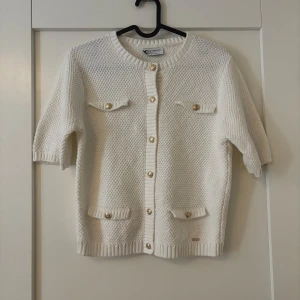 Vit stickad kofta med guldknappar - Supersöt vit stickad kofta från Kelly & White med kort ärm och snygga guldiga knappar framtill. Koftan har fyra fickor med lock och ribbade kanter. Perfekt att slänga över en topp för en chic look.