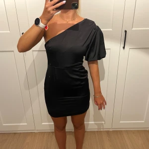 Svart one shoulder klänning Boohoo - Snygg svart kortklänning från Boohoo med one shoulder-design och puffärm. Klänningen har en tight passform och är tillverkad i ett glansigt material som ger en lyxig känsla. Perfekt för dig som vill sticka ut med en trendig och elegant look.