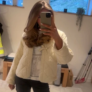 Quiltad vit jacka med rosett - Supersöt quiltad jacka i vitt med en fin rosettdetalj framtill. Jackan har en croppad passform och långärmade ärmar. Perfekt för dig som gillar en clean och trendig look.